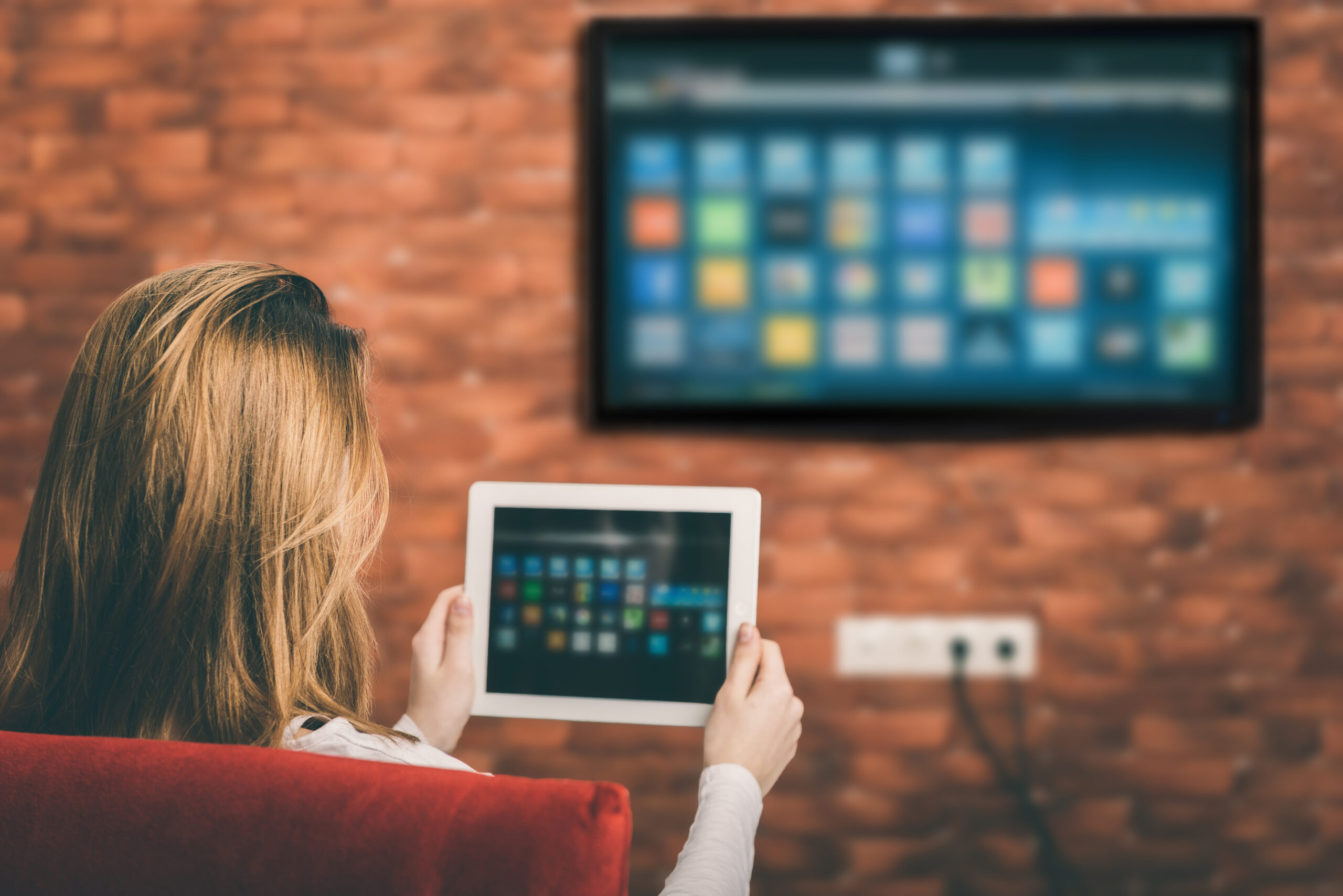 Decorative image for how CTV is transforming user engagement. This image shows a woman sitting in front of a television that shows options for different apps she could watch. The woman is also holding a tablet that also shows options for apps she can use to watch content on.